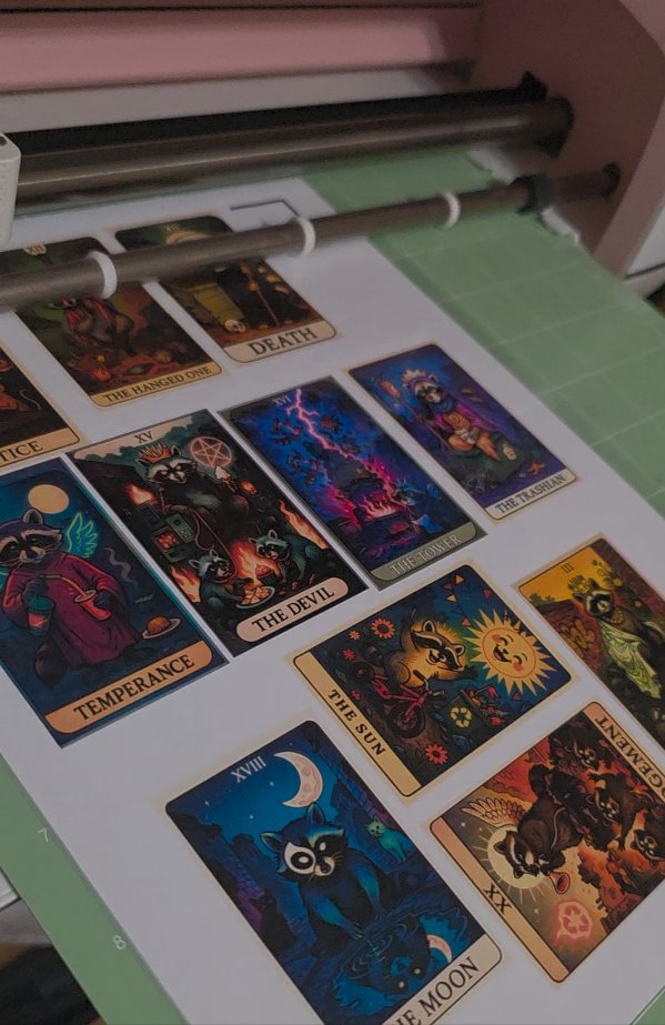 Printed Oracle of the Moonlit Dumpster cards laid out on a cutting mat mid-cut, including The Hanged One, Death, The Devil, The Tower, Temperance, The Sun, and The Moon
