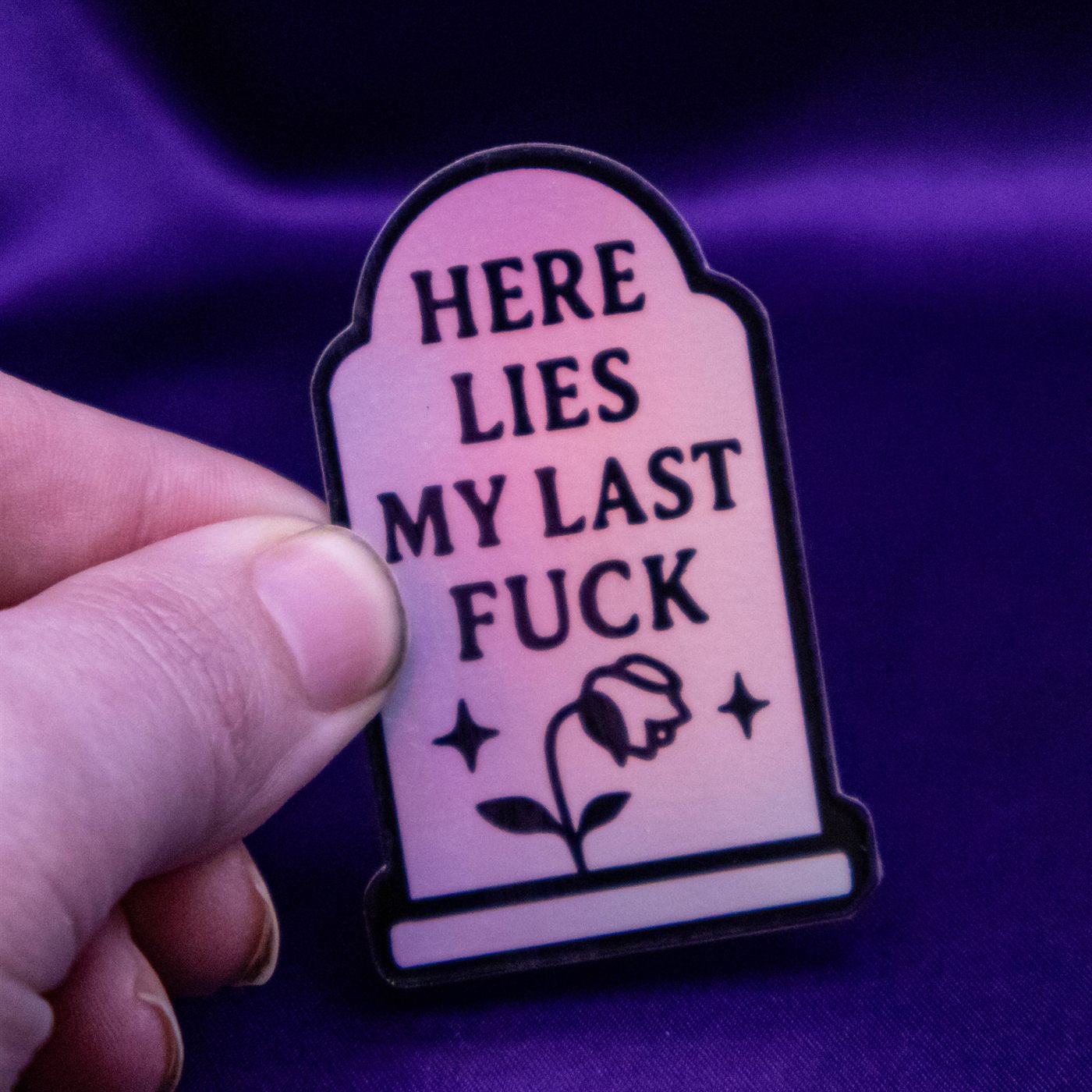 A laminated holographic Neurospice Design Shop sticker reading Here Lies My Last Fuck, dark humor permission slip