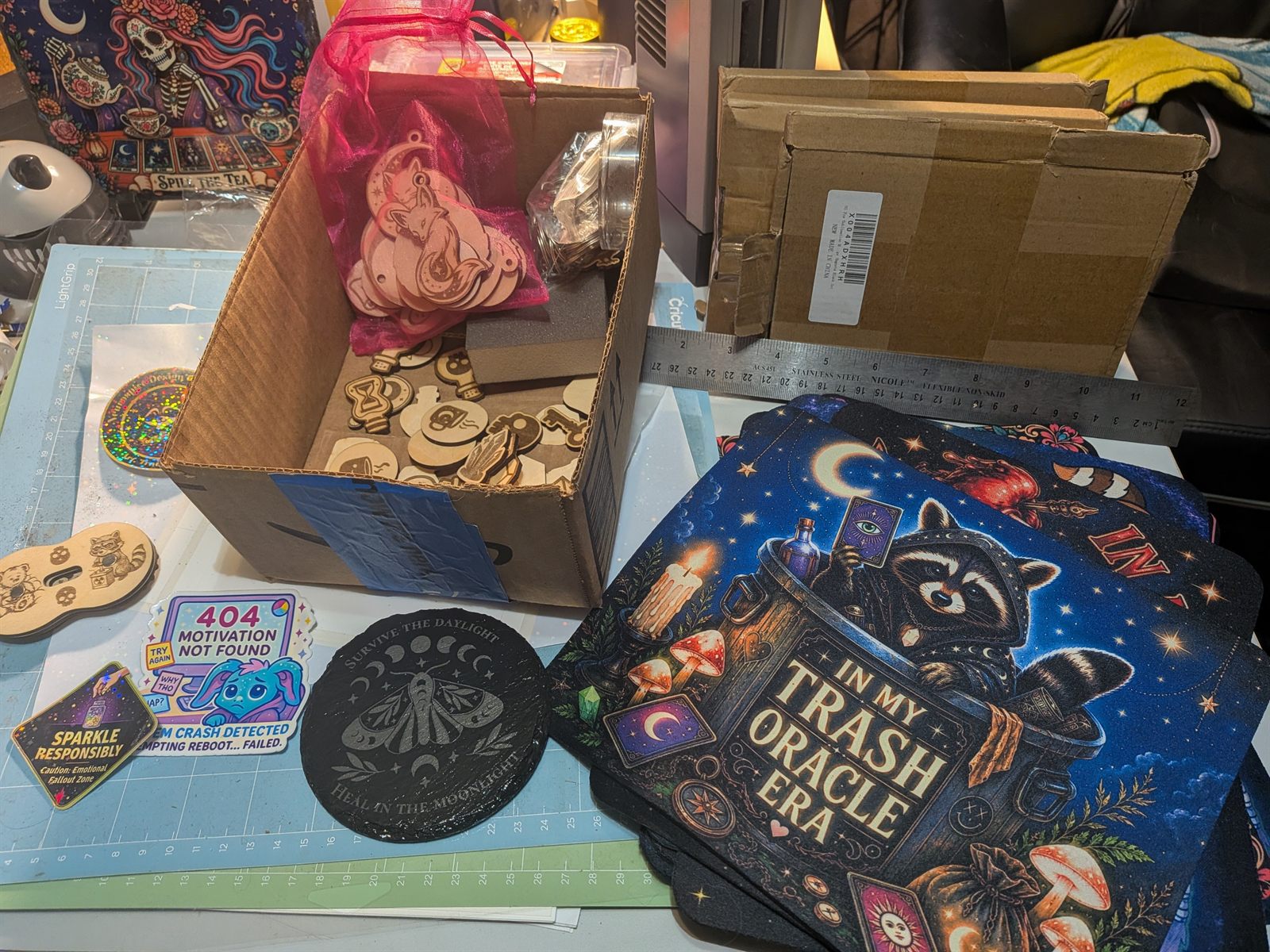 The Neurospice Design Shop workspace, a box of laser-cut wooden charms beside a Trash Oracle mousepad and stickers