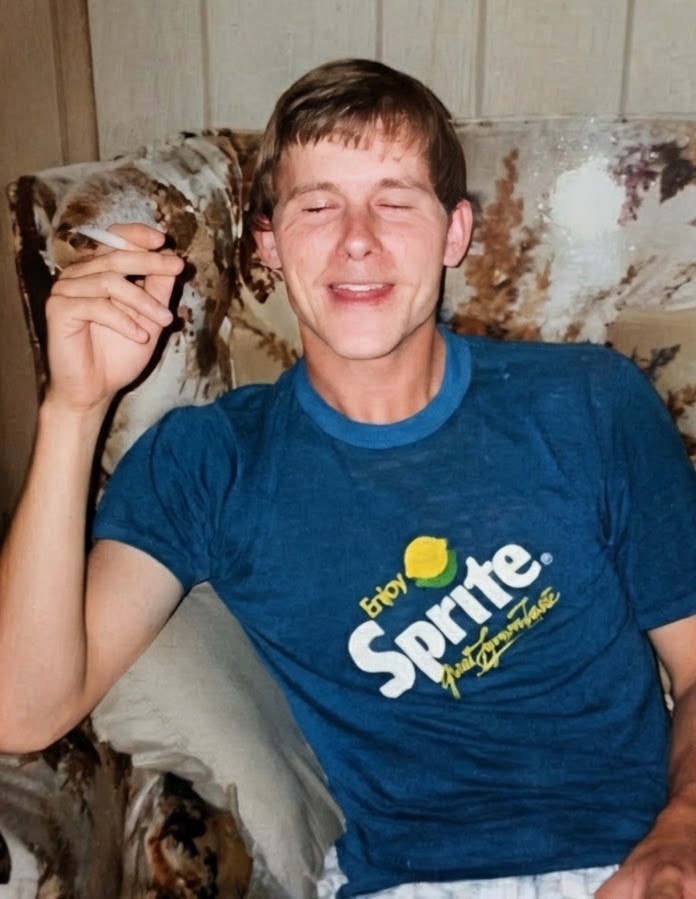 Laura's dad as a young man, laughing with his eyes squeezed shut, sitting in a floral armchair in a blue Sprite ringer tee
