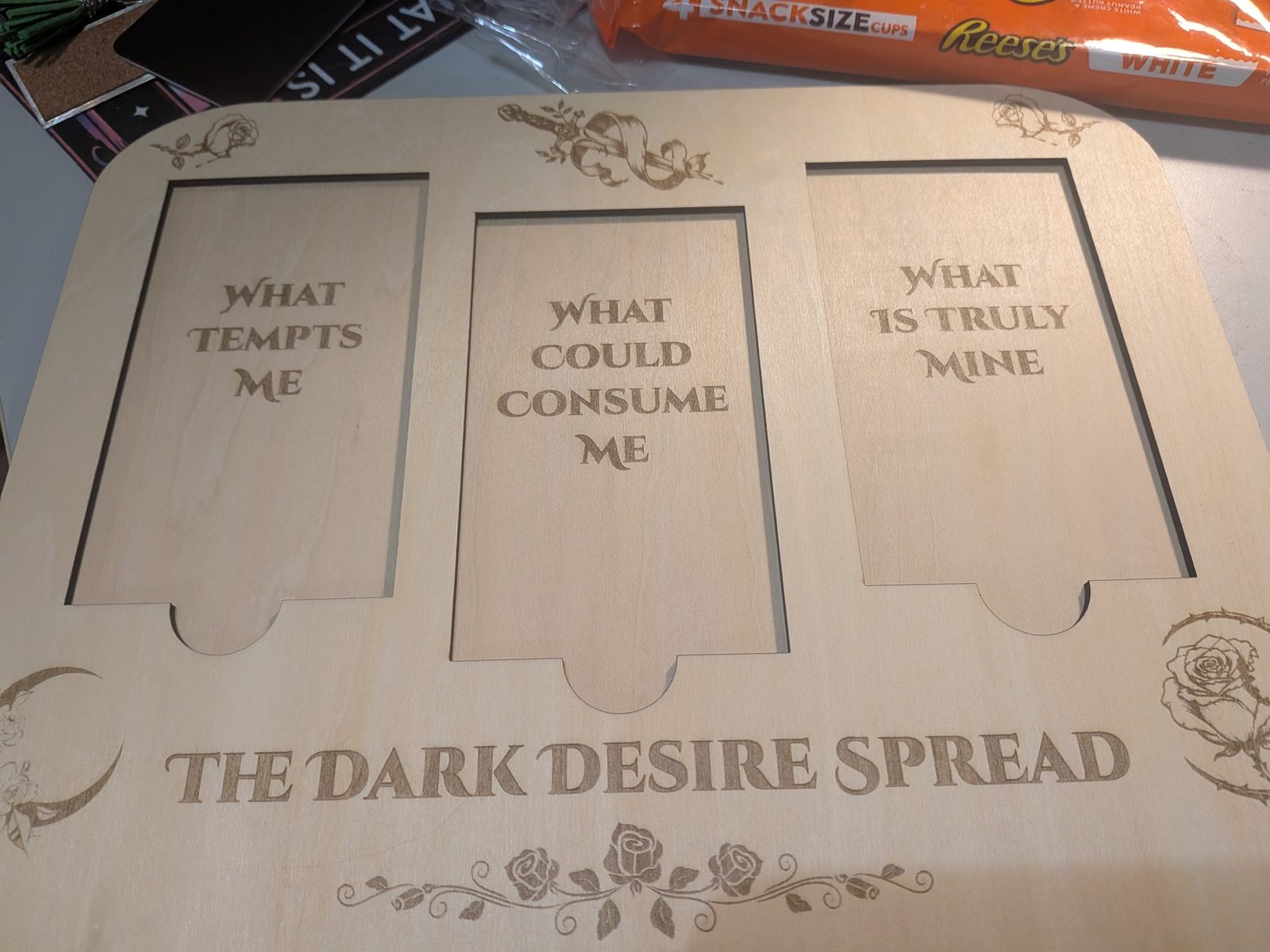 A laser-engraved wooden tarot spread board reading The Dark Desire Spread, on a workbench with a bag of Reese's cups behind it