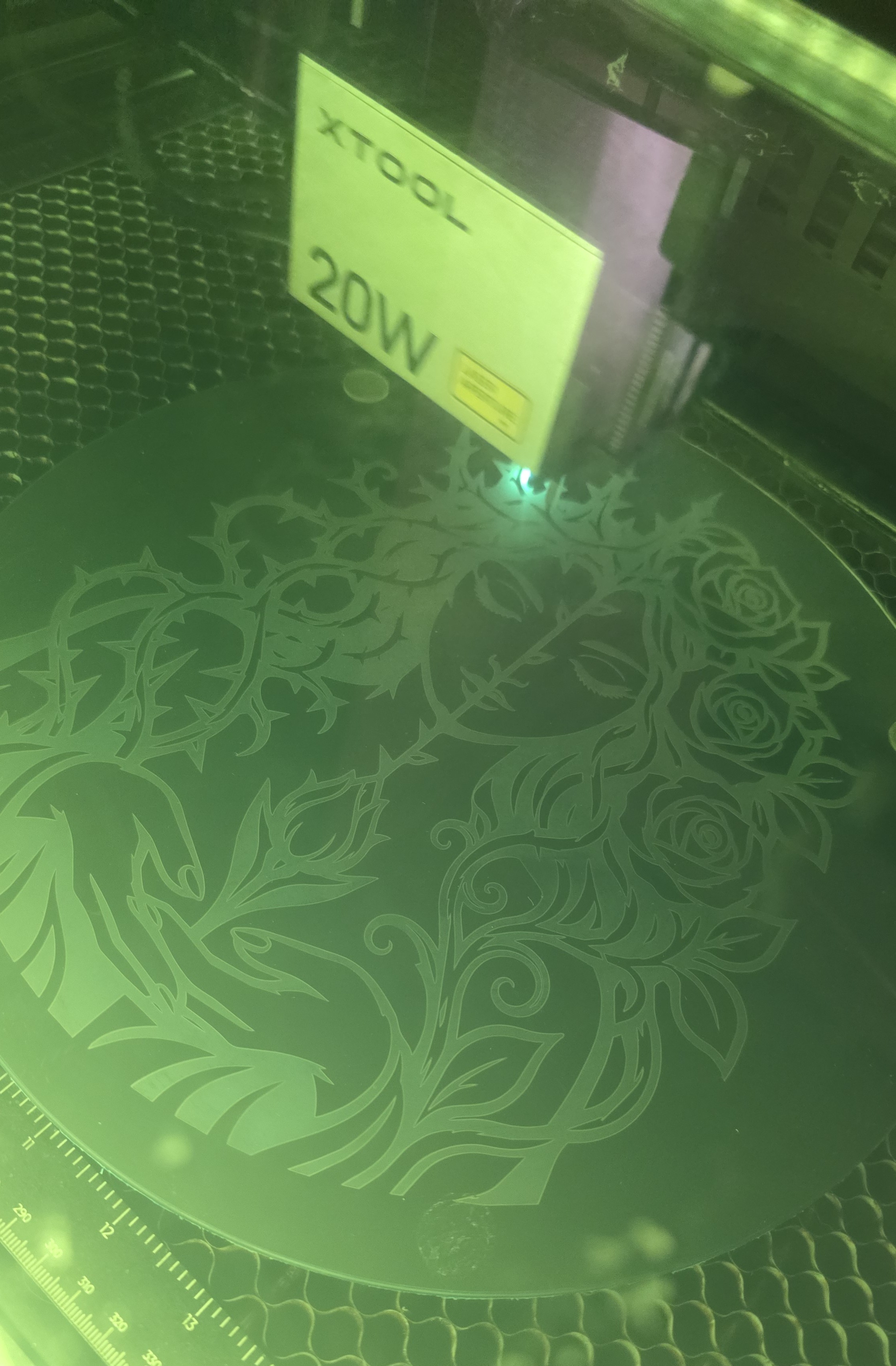 An xTool 20W laser engraving an intricate floral design on a round piece of wood, viewed through the green safety window